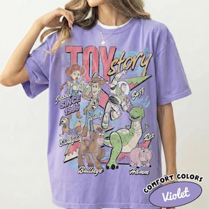May include: A light purple short-sleeve t-shirt featuring a graphic print of Toy Story characters including Woody, Buzz Lightyear, Jessie, Slinky Dog, Bullseye, Rex, and Hamm. The text reads TOY story, Jessie SINCE 1995, Woody, Buzz, Slinky Dog, Bullseye, Rex, and Hamm.