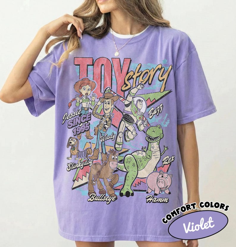May include: A light purple short-sleeve t-shirt featuring a graphic print of Toy Story characters including Woody, Buzz Lightyear, Jessie, Slinky Dog, Bullseye, Rex, and Hamm. The text reads TOY story, Jessie SINCE 1995, Woody, Buzz, Slinky Dog, Bullseye, Rex, and Hamm.