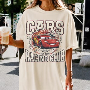 May include: Cream-colored t-shirt featuring a graphic of a red race car with the text "CARS RACING CLUB EST. 2006". The design includes a cartoon car breaking through a wall, with a vintage, distressed aesthetic.