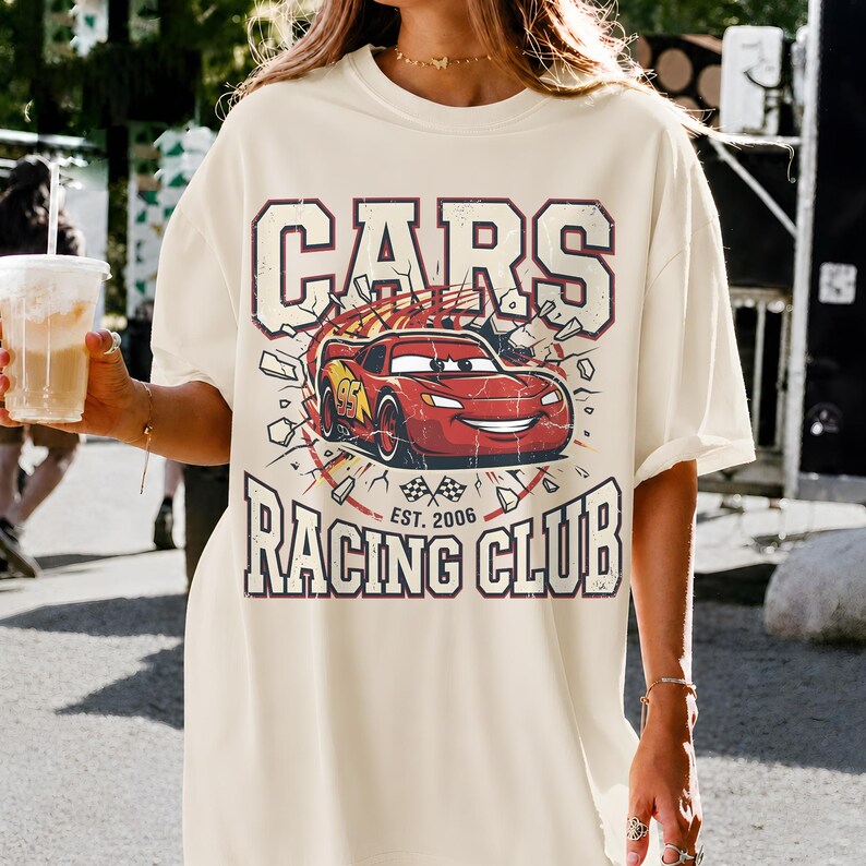 May include: Cream-colored t-shirt featuring a graphic of a red race car with the text "CARS RACING CLUB EST. 2006". The design includes a cartoon car breaking through a wall, with a vintage, distressed aesthetic.