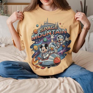 May include: Mustard yellow t-shirt featuring a colorful graphic of Mickey Mouse, Donald Duck, and Goofy in space suits on a rocket ship. The words "SPACE MOUNTAIN" are printed above the characters in a retro font, with planets and stars in the background.