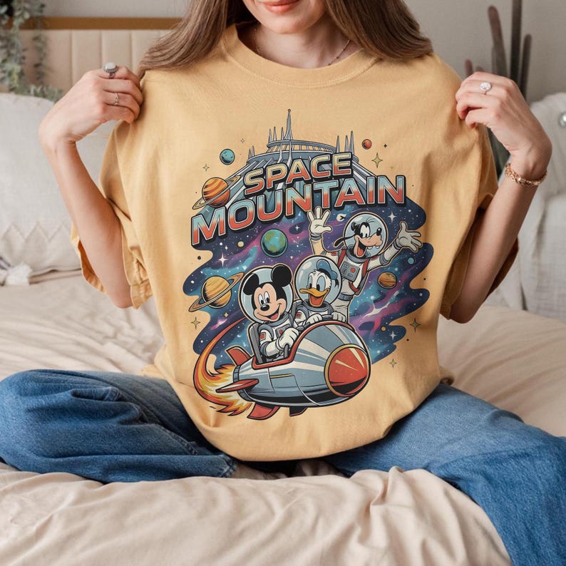 May include: Mustard yellow t-shirt featuring a colorful graphic of Mickey Mouse, Donald Duck, and Goofy in space suits on a rocket ship. The words "SPACE MOUNTAIN" are printed above the characters in a retro font, with planets and stars in the background.