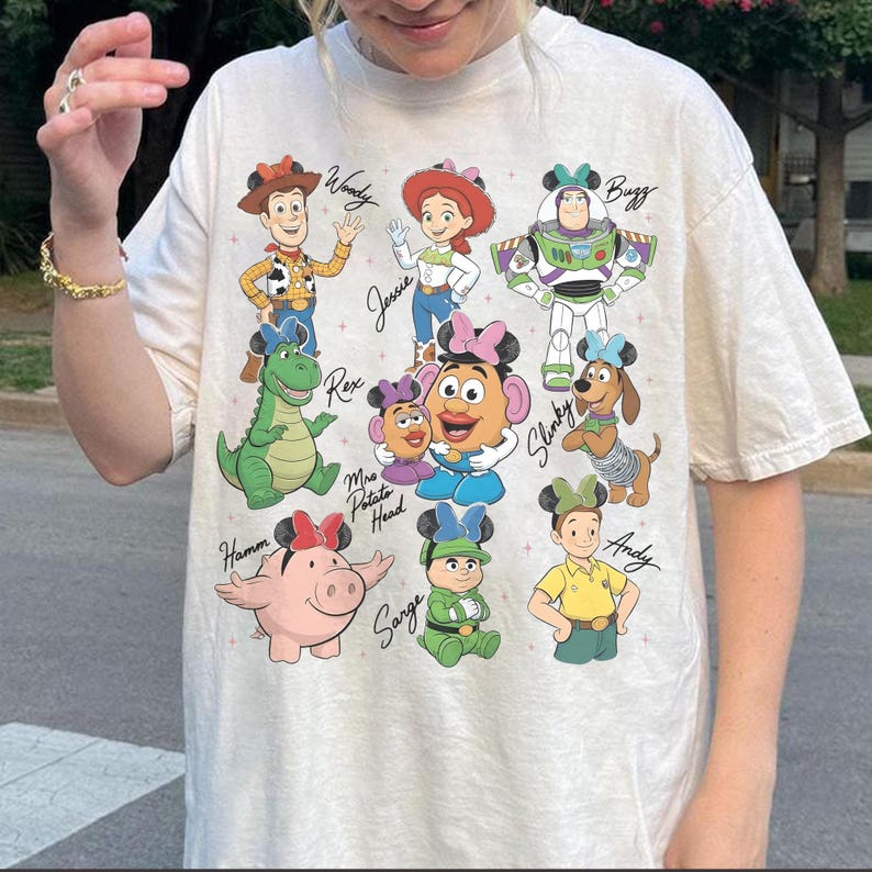 May include: Cream-colored t-shirt featuring a colorful graphic of Toy Story characters, including Woody, Jessie, Buzz Lightyear, and others. Each character is depicted with a Mickey Mouse ear headband. The names of the characters are written in a handwritten font.