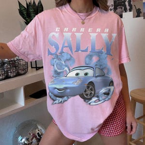 May include: A light pink t-shirt featuring a graphic of a blue cartoon car with the name "SALLY" in large, stylized letters above it. The word "CARRERA" is printed above "SALLY". The shirt has short sleeves.