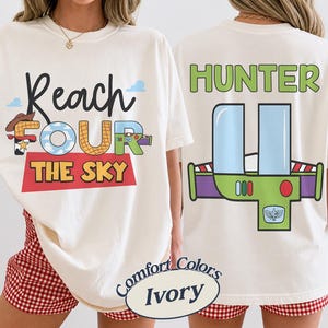 以下が含まれることがあります： 黒、青、赤、黄色の「Reach FOUR the SKY」の文字が入ったアイボリー色のTシャツ。背中には、緑、紫、青のグラフィックの上に緑色の「HUNTER」という名前がプリントされています。このシャツはComfort Colorsコレクションの一部です。