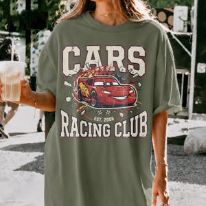 May include: Olive green t-shirt with a graphic of a red race car with the text "CARS RACING CLUB EST. 2006". The car is breaking through a wall. The shirt has a vintage, distressed look.