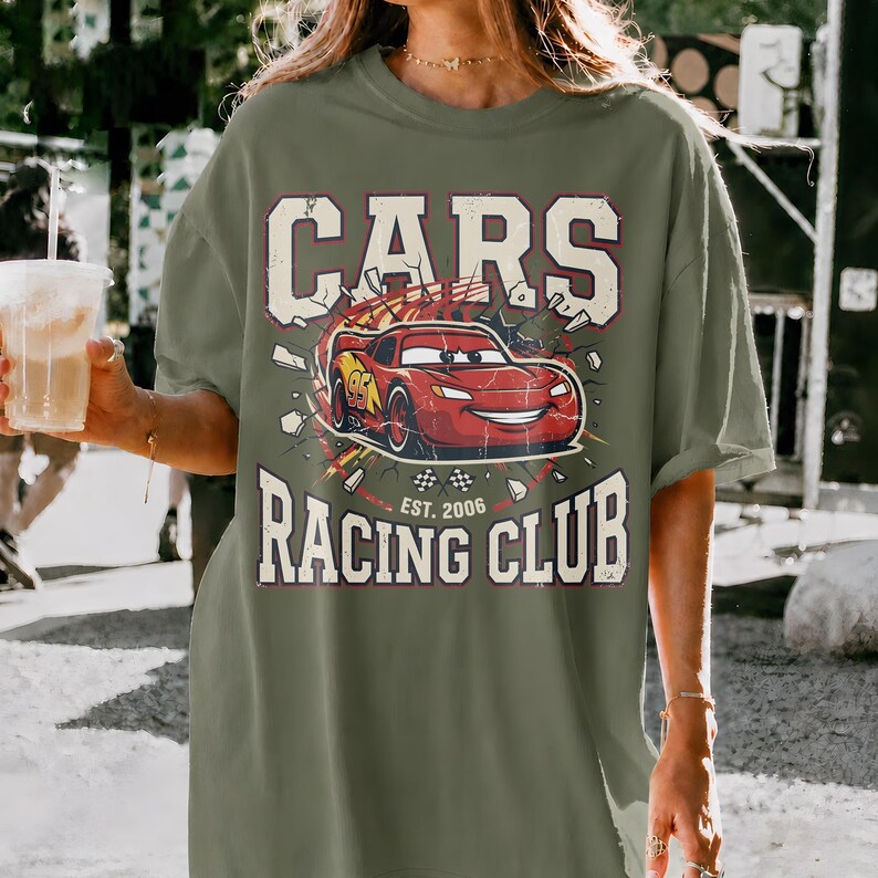 May include: Olive green t-shirt with a graphic of a red race car with the text "CARS RACING CLUB EST. 2006". The car is breaking through a wall. The shirt has a vintage, distressed look.