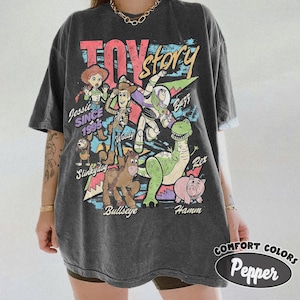 May include: A charcoal gray short-sleeve t-shirt featuring a graphic print of Toy Story characters including Woody, Buzz Lightyear, Jessie, Rex, Hamm, Bullseye, and Slinky Dog. The text reads Toy Story, Jessie Since 1995, Woody, Buzz, Rex, Hamm, and Bullseye.