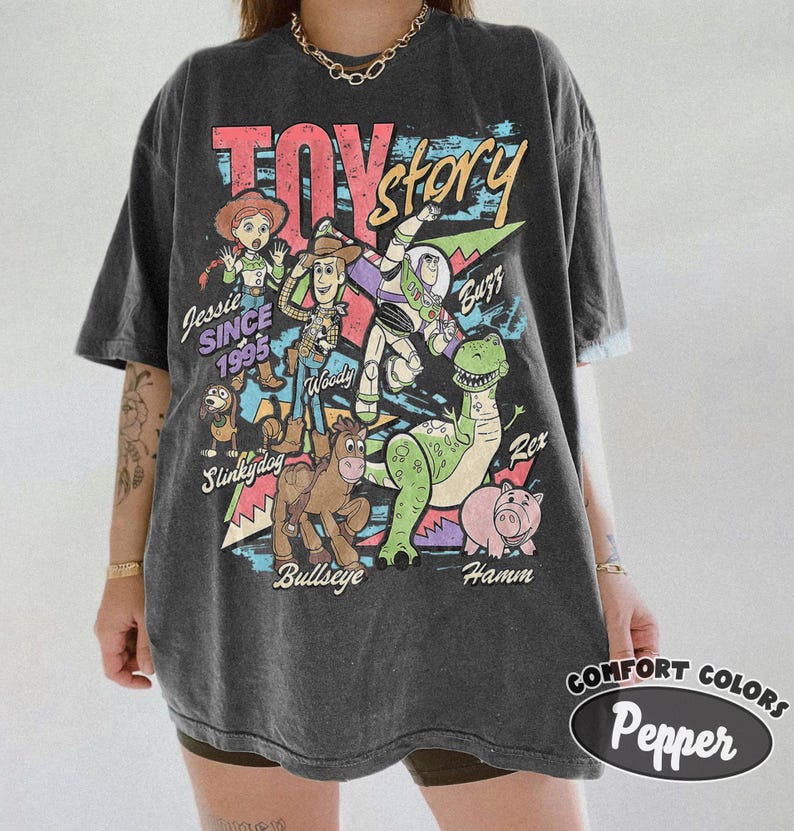 May include: A charcoal gray short-sleeve t-shirt featuring a graphic print of Toy Story characters including Woody, Buzz Lightyear, Jessie, Rex, Hamm, Bullseye, and Slinky Dog. The text reads Toy Story, Jessie Since 1995, Woody, Buzz, Rex, Hamm, and Bullseye.