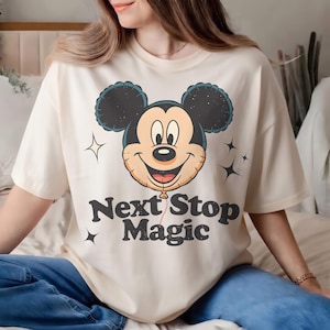 May include: Cream-colored t-shirt featuring a smiling Mickey Mouse balloon graphic with the text "Next Stop Magic" in black. The design includes star accents. A casual, comfortable top.