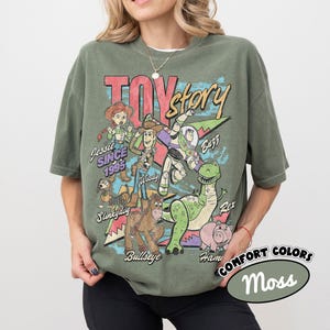 May include: An olive green short-sleeve t-shirt featuring a vintage-style graphic of Toy Story characters including Woody, Buzz Lightyear, Jessie, Slinky Dog, Bullseye, Rex, and Hamm. The text reads Toy Story, Jessie SINCE 1995, Buzz, Slinky Dog, Bullseye, Rex, and Hamm.