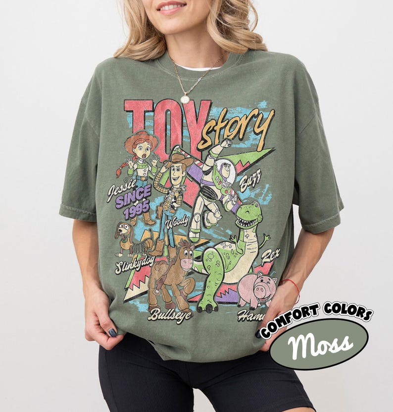 May include: An olive green short-sleeve t-shirt featuring a vintage-style graphic of Toy Story characters including Woody, Buzz Lightyear, Jessie, Slinky Dog, Bullseye, Rex, and Hamm. The text reads Toy Story, Jessie SINCE 1995, Buzz, Slinky Dog, Bullseye, Rex, and Hamm.