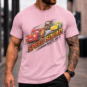 Op de afbeelding: Een lichtroze t-shirt met de tekst "Speed Squad Route 66 Radiator Springs" en illustraties van drie cartoonauto's. De auto's zijn rood, bruin en geel, met een racethema.
