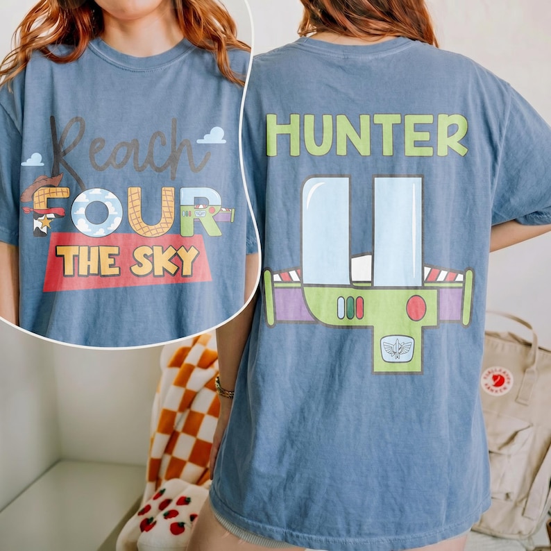 以下が含まれることがあります： 前面に「Reach for the Sky」の文字と玩具箱のグラフィックが入った青いTシャツ。背中には「HUNTER」という名前とバズ・ライトイヤーのデザインが施されています。快適でカジュアルなスタイルです。