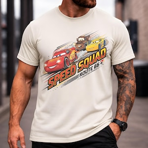 Op de afbeelding: Beige T-shirt met een afbeelding van Bliksem McQueen, Takel en een gele auto uit de film Cars. De tekst "Speed Squad Route 66 Radiator Springs" is onder de auto's gedrukt in een dynamisch ontwerp.