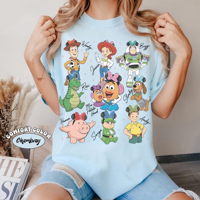 May include: Light blue t-shirt featuring cartoon characters Woody, Jessie, Buzz, Rex, Mr. Potato Head, Slinky, Hamm, Sarge, and Andy. The shirt has the words "COMFORT COLOR" and "Chambray" printed on it.