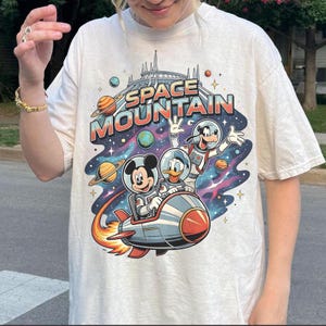 May include: Off-white t-shirt featuring a colorful graphic of Mickey Mouse, Donald Duck, and Goofy in space suits, riding a rocket ship. The text "SPACE MOUNTAIN" is above them, with planets and stars in the background.