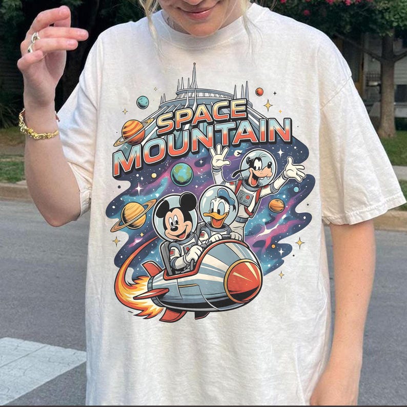 May include: Off-white t-shirt featuring a colorful graphic of Mickey Mouse, Donald Duck, and Goofy in space suits, riding a rocket ship. The text "SPACE MOUNTAIN" is above them, with planets and stars in the background.