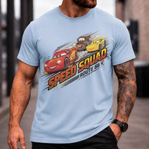 Op de afbeelding: Lichtblauw T-shirt met de tekst "Speed Squad Route 66 Radiator Springs" en illustraties van drie cartoonauto's. De afbeelding bevat een rode auto, een bruine truck en een gele auto.