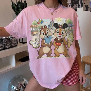 May include: Pink t-shirt featuring a cartoon graphic of Chip and Dale holding treats in front of a park scene with a castle, food cart, and balloons. The shirt has a crew neck and short sleeves.