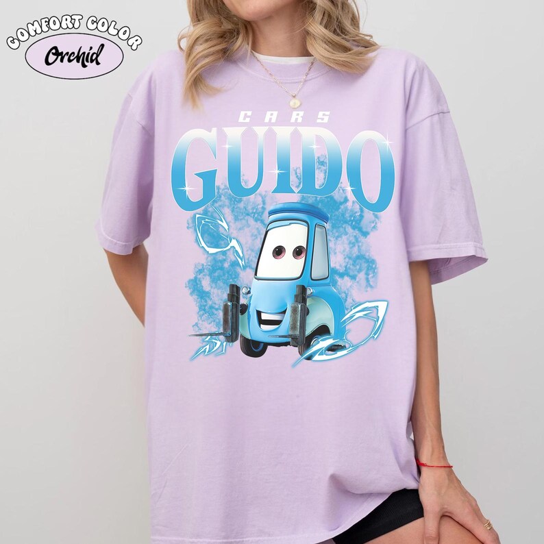 May include: A light purple Comfort Colors t-shirt featuring the word "GUIDO" in large, light blue letters with a cartoon image of Guido from the movie Cars. The shirt also has the word "CARS" above the name and the word "Orchid" in a circle.