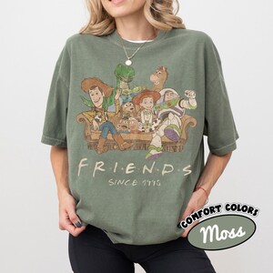 May include: A moss green short-sleeve t-shirt featuring a graphic print of Toy Story characters sitting on a couch. The text "FRIENDS" and "SINCE 1995" is printed below the characters. The shirt has a crew neckline and a relaxed, loose fit.
