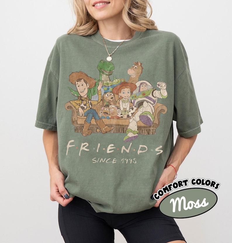 May include: A moss green short-sleeve t-shirt featuring a graphic print of Toy Story characters sitting on a couch. The text "FRIENDS" and "SINCE 1995" is printed below the characters. The shirt has a crew neckline and a relaxed, loose fit.
