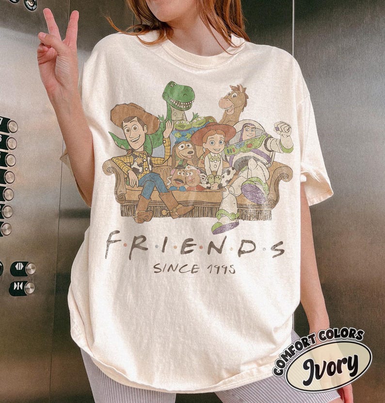 May include: An ivory t-shirt featuring a graphic print of Toy Story characters sitting on a couch with the text "F.R.I.E.N.D.S SINCE 1995" printed below. The characters include Woody, Buzz Lightyear, Jessie, Rex, Slinky Dog, Bullseye, and Mr. Potato Head.