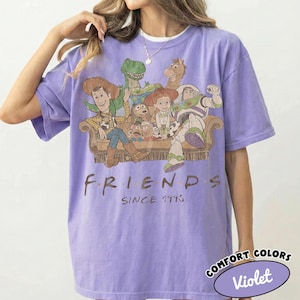 May include: A light purple short-sleeve t-shirt featuring a graphic print of Toy Story characters sitting on a couch. Below the characters, the text reads FRIENDS SINCE 1995. A small oval label in the corner displays the text COMFORT COLORS Violet.