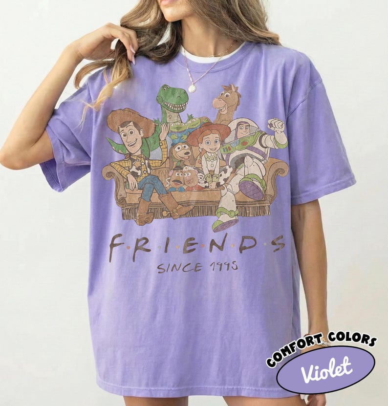 May include: A light purple short-sleeve t-shirt featuring a graphic print of Toy Story characters sitting on a couch. Below the characters, the text reads FRIENDS SINCE 1995. A small oval label in the corner displays the text COMFORT COLORS Violet.