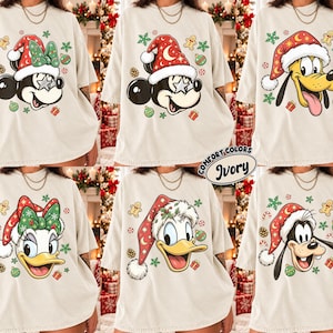 Könnte beinhalten: Ein elfenbeinfarbenes T-Shirt mit einem Raster aus sechs Zeichentrickfiguren, die rot-weiße Weihnachtsmützen tragen. Die Figuren umfassen Minnie Maus, Mickey Maus, Pluto, Daisy Duck, Donald Duck und Goofy, umgeben von Lebkuchenmännern.