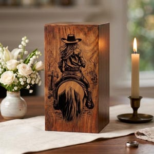 Op de afbeelding: Een bruine houten kist met een gedetailleerde gravure van een persoon te paard, met een cowboyhoed. De kist staat op een houten tafel. Een brandende kaars in een messing houder staat ernaast.