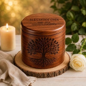 May include: A wooden urn with a tree of life design, resting on a wooden slice. The top of the urn is engraved with the name "ALEXANDER CHEN", dates "1968-2026", and the phrase "FOREVER IN OUR HEARTS". A lit candle and a white rose are in the background.