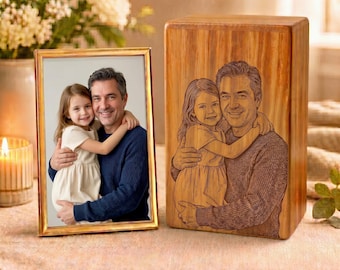 May include: A rectangular wooden cremation urn featuring a laser-engraved portrait of a child being held by an adult, displayed next to a matching framed photograph of the same subjects. The urn has a natural wood grain finish and vertical orientation.
