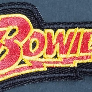 Puede incluir: Un parche bordado que presenta la palabra Bowie en letras rojas con un contorno amarillo. El texto se encuentra sobre un fondo negro con una forma estilizada y dentada que se extiende hacia la izquierda, creando un elemento de diseño angular.