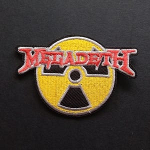 May include: A circular embroidered patch featuring a yellow and black radiation symbol. The word Megadeth is stitched in red lettering with a white outline across the centre of the symbol. The patch has a black border and detailed stitching.