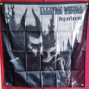 Pode incluir: Um estandarte de parede quadrado de tecido preto e branco com o texto Electric Wizard e Dopethrone no canto superior direito. O gráfico retrata uma figura escura e com chifres com uma estética esfumaçada e borrada, com ilhós metálicos.