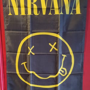 May include: A rectangular black fabric wall banner featuring the yellow text NIRVANA at the top and a large yellow smiley face graphic below. The smiley face has X-shaped eyes and a wavy mouth with a tongue sticking out. Metal grommets are in each corner.