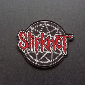 May include: A circular embroidered patch featuring a black background with a white geometric star design. The word Slipknot is stitched in a jagged, red font across the center of the patch, with a thick black border outlining the entire shape.