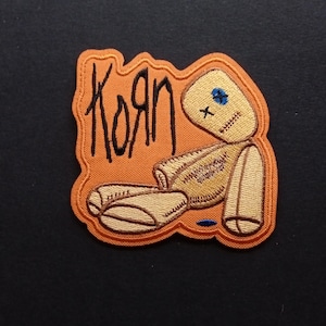 May include: An embroidered patch featuring a tan rag doll with a blue stitched eye and a stitched mouth, positioned against an orange background. The text KoRn is embroidered in black on the left side of the patch, which has a scalloped orange border.