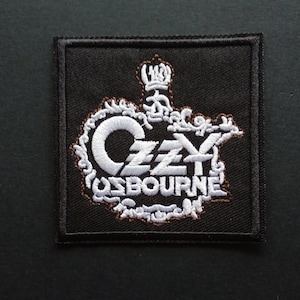 May include: A square black embroidered patch featuring the white text Ozzy Osbourne in a stylized font. The design includes a white crown icon above the name, surrounded by intricate white scrollwork and thin brown stitching outlines.