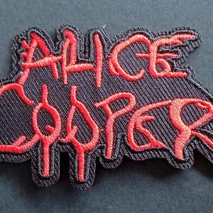 Peut inclure: Un écusson brodé noir présentant le texte Alice Cooper dans une police rouge stylisée. Les lettres sont cousues avec du fil rouge sur une base en tissu noir, créant un contraste marqué. L'écusson a une forme distincte, dentelée et irrégulière.