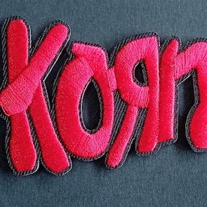 Peut inclure: Un écusson brodé représentant le mot Korn en fil rouge vif avec une bordure noire épaisse. Le lettrage est stylisé avec une police dentelée distincte où la lettre R est inversée, créant un design graphique audacieux pour application textile.