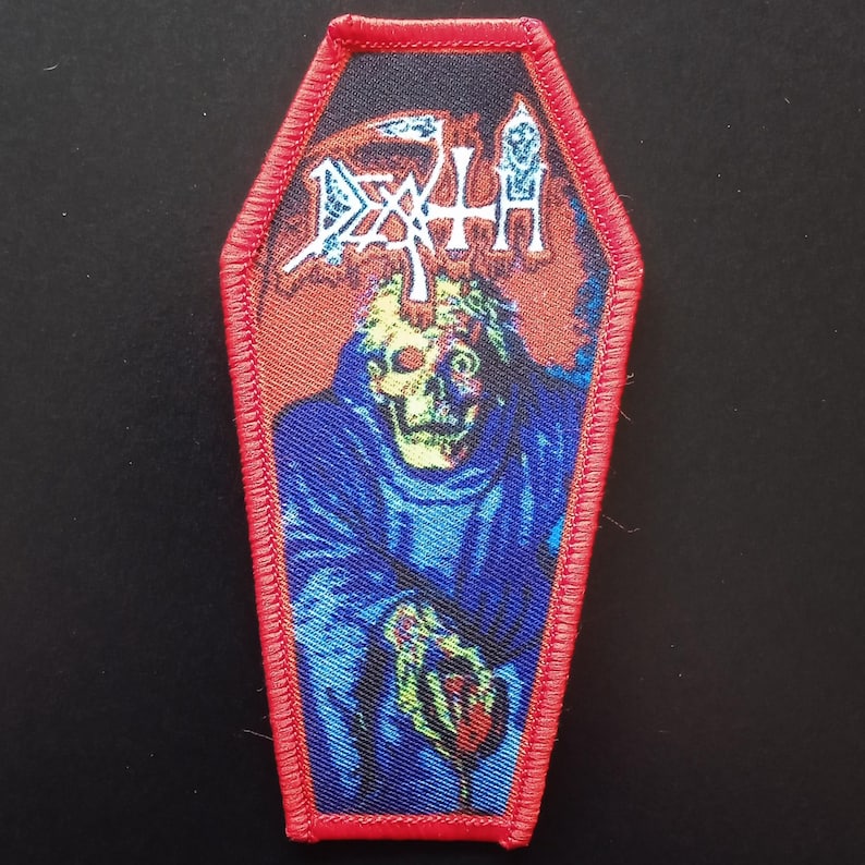 May include: A coffin-shaped embroidered patch with a red stitched border. The design features a graphic of a skeletal figure in a blue robe with the word DEATH printed in white stylized lettering at the top against a dark background.