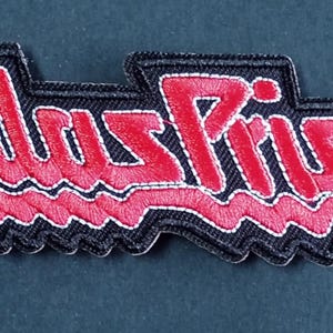 Op de afbeelding: Een rechthoekige geborduurde patch met de tekst Judas Priest in een gestileerd rood lettertype met een dikke zwarte rand. De letters hebben een opvallend grillig, hoekig ontwerp met witte stikselaccenten rond de rode tekens.