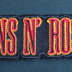Puede incluir: Un parche bordado rectangular que presenta el texto GUNS N' ROSES en letras negritas rojas y amarillas. Las letras están delineadas con hilo negro sobre un fondo de tela negra, con un borde grueso cosido en negro que rodea el diseño.