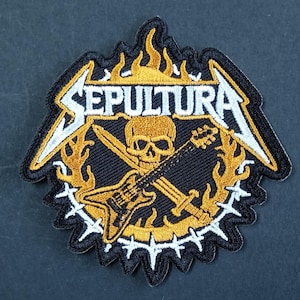 May include: An embroidered patch featuring the word SEPULTURA in white lettering above a gold skull, a black electric guitar, and a sword. The design is surrounded by gold flames and a white barbed wire border on a black background.