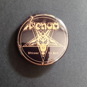 Peut inclure: Bouton rond noir avec lettrage et motif dorés. Le bouton présente le mot "Venom", un pentagramme avec une tête de chèvre et les mots "Welcome To Hell".