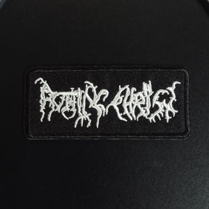 May include: A rectangular embroidered patch featuring a black background with white text that reads Rotting Christ in a stylized, jagged, and intricate font. The patch has a stitched border and is designed for application to fabric items.