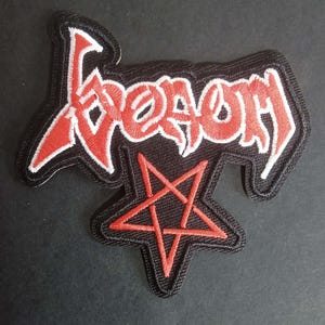 Puede incluir: Parche bordado con la palabra "Venom" en letras góticas rojas con contorno blanco, sobre un fondo negro. Un pentagrama rojo está debajo. El parche tiene un borde negro.